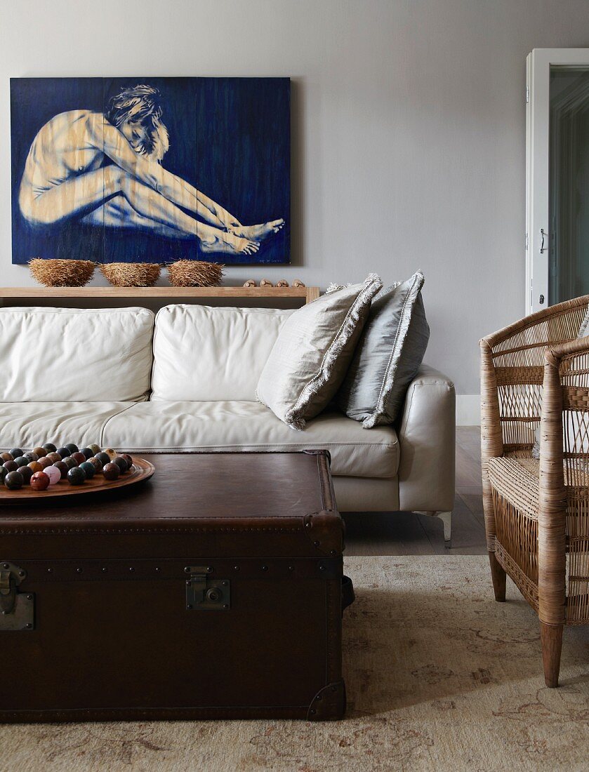 White living room couch below nude on wall; antique leather trunk as coffee table and simple wicker armchair in foreground