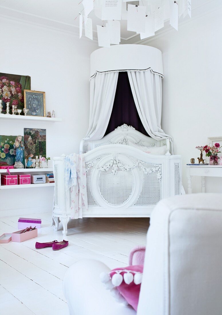 Romantic, white bed with round canopy on white wooden floor; various artworks leaning against wall on floating shelves