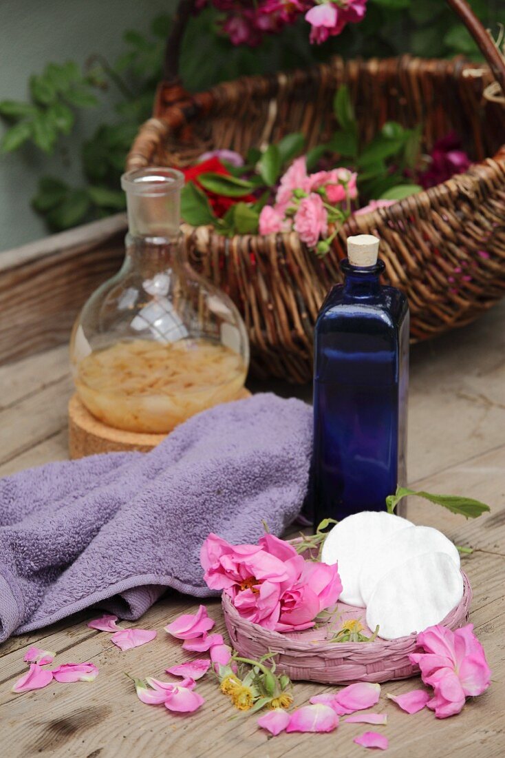An arrangement of home-made rosewater in a blue bottle next to a towel, roses and rose petals