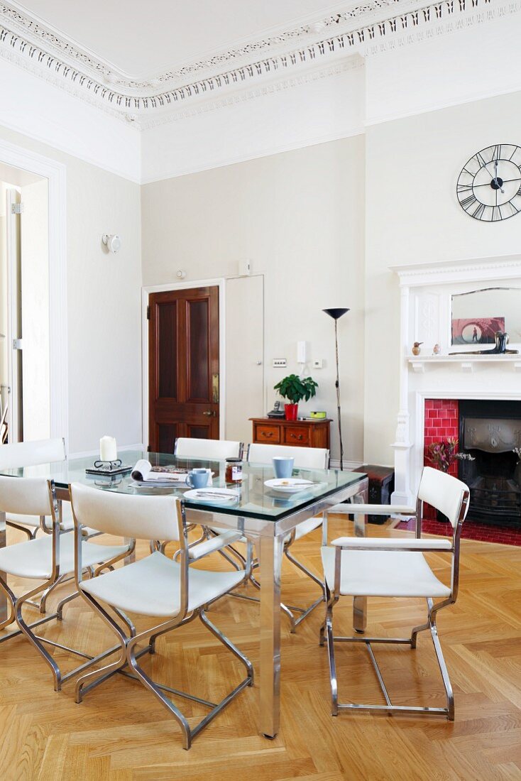 Dining table with glass top and white leather chairs in grand interior with open fireplace in background and stucco frieze on ceiling