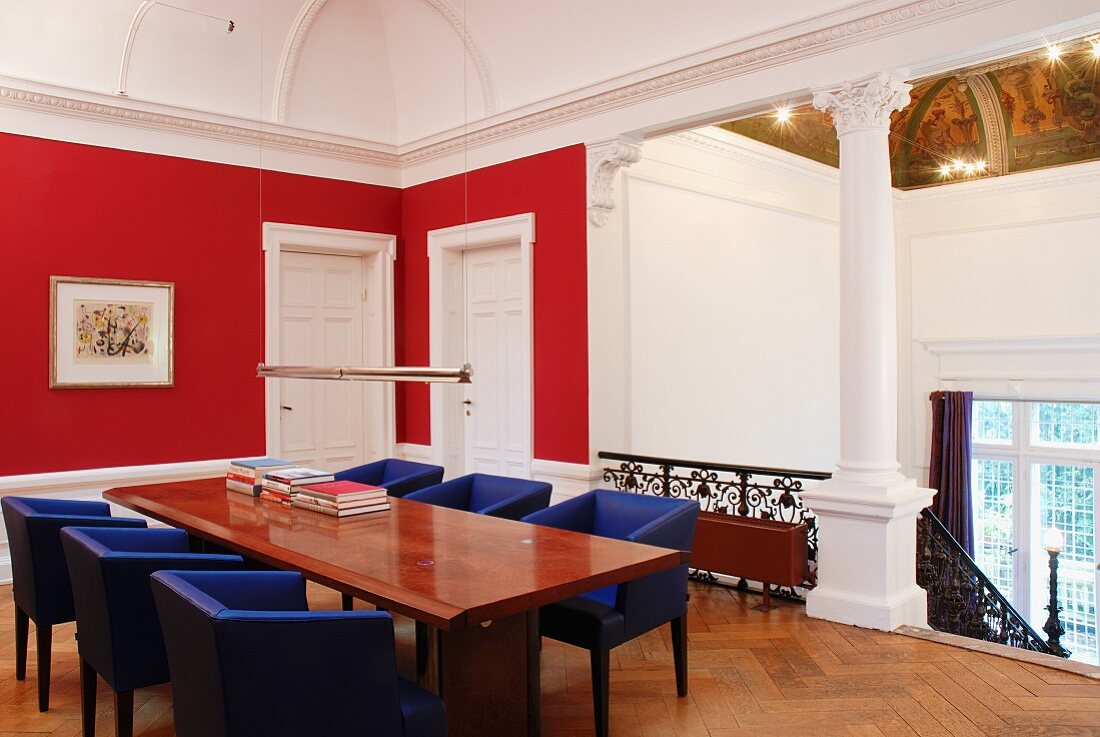Landing with red-painted walls in grand villa - table and blue-covered armchairs at head of stairs with Corinthian columns below stucco picture rail
