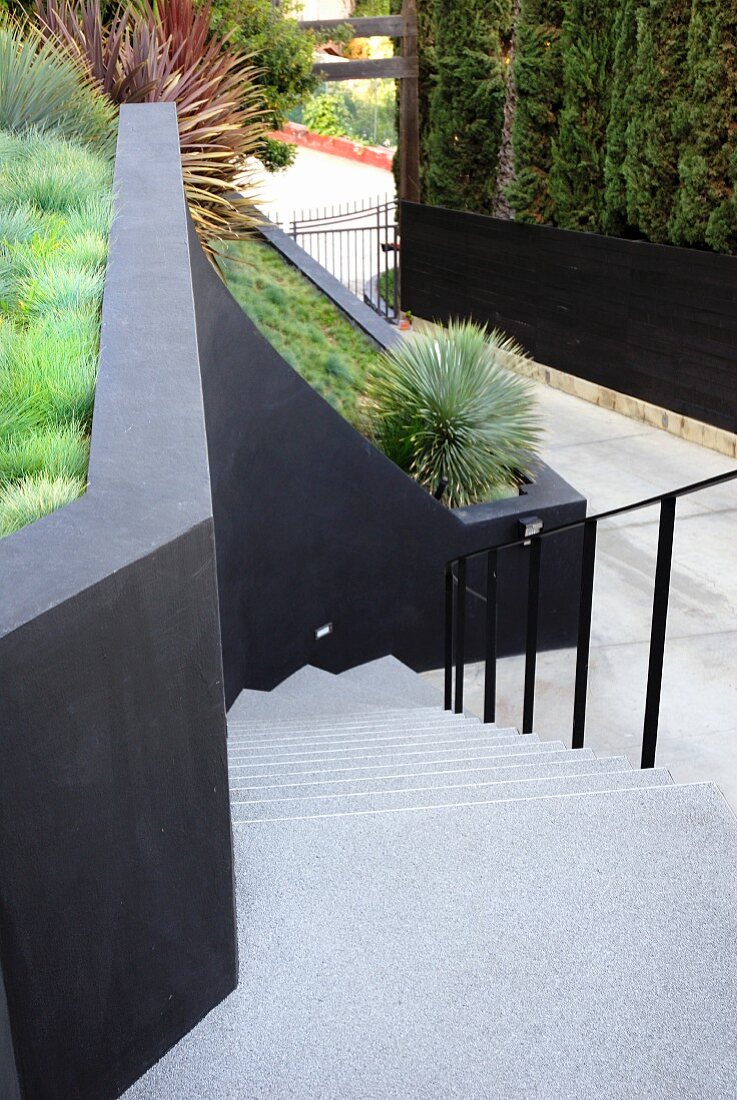 Blick von oben auf Treppe aus hellem Beton und schwarzes Metallgeländer gegenüber schwarzer Wand, dahinter terrassierter Garten