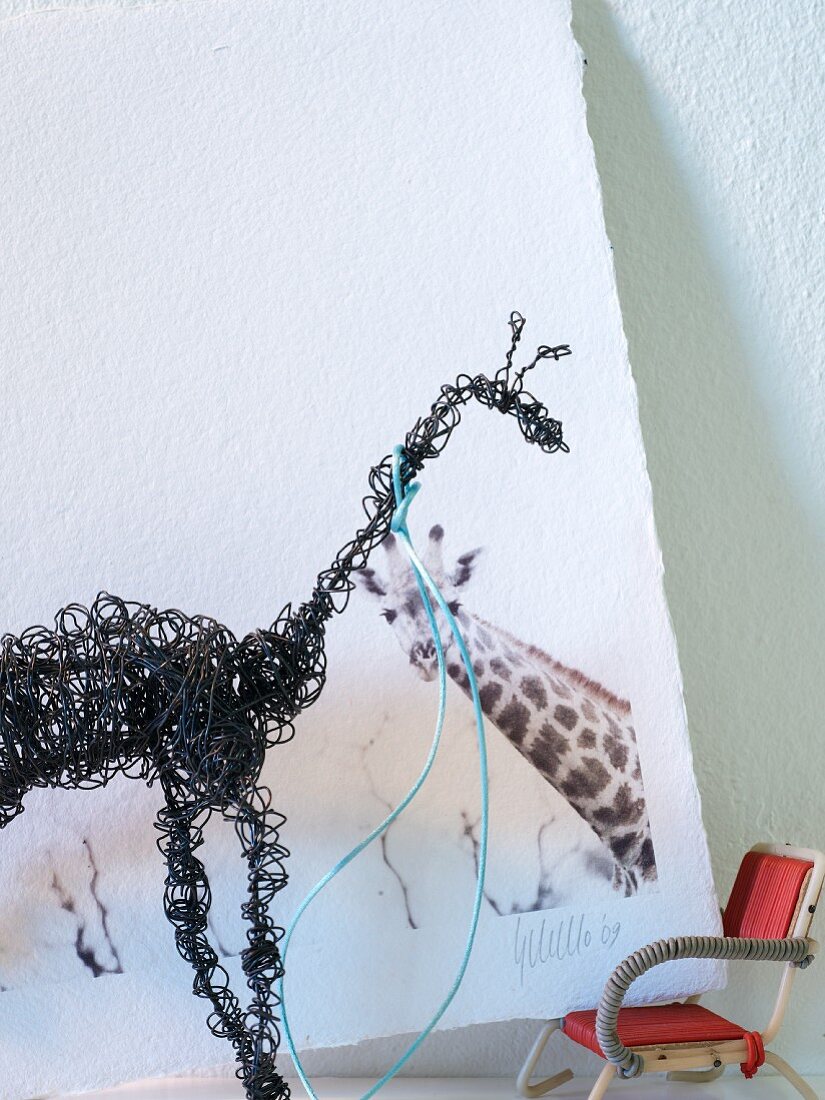Black, wire giraffe sculpture in front of picture of giraffe on white background