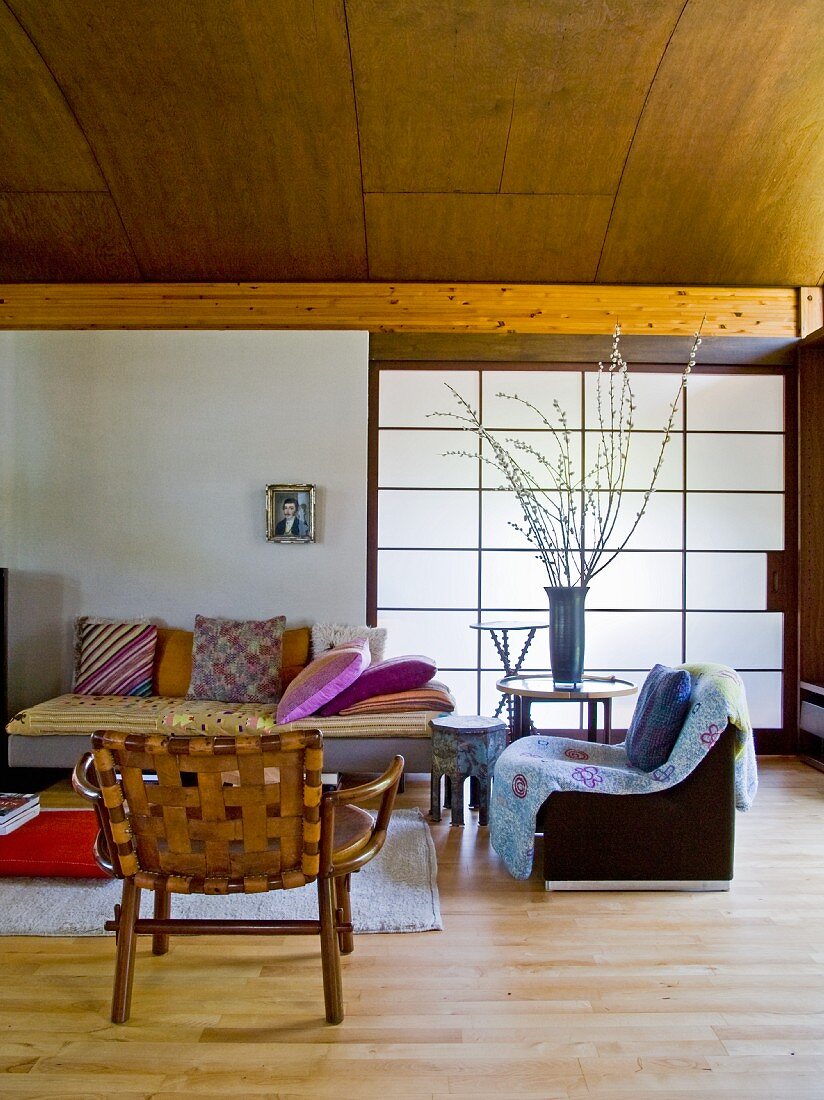 Holzstuhl und Sessel neben gemütlichem Sofa mit Kissen vor Schiebeelement mit japanischem Papier bespannt in modernem Wohnraum mit holzverkleideter Tonnendecke