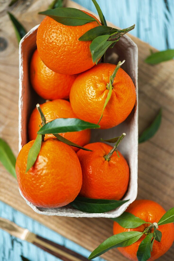 Mehrere Mandarinen mit Blättern im Pappschälchen auf Schneidebrett