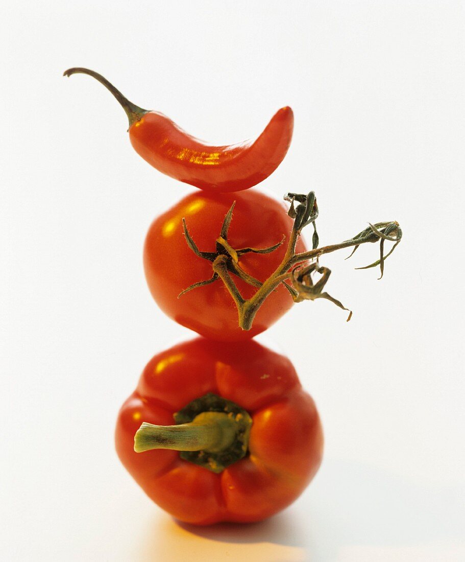 A still life featuring a pepper, a tomato and a chilli, stacked one on top of the other