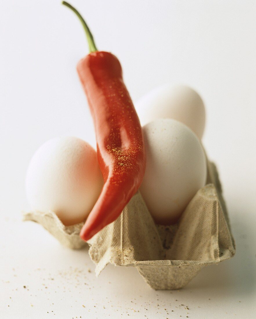 A still life featuring a chilli pepper and white hen's eggs in an eggbox