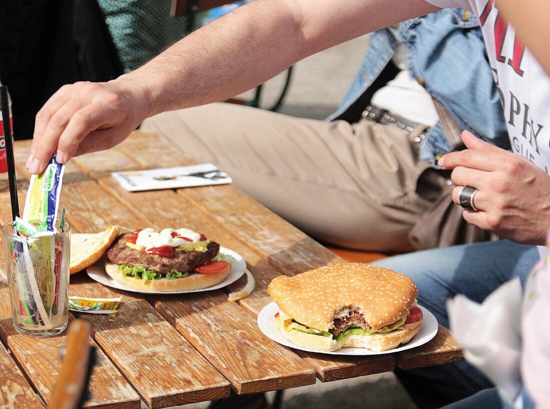 Burgers on a table in a beer garden in the sunshine; a man's hand reaching for a sachet of mayonnaise