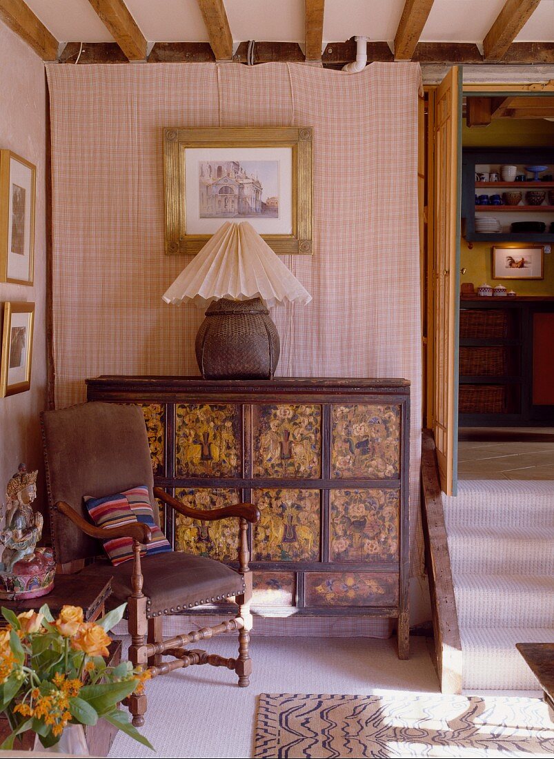 The corner of a room in an English country house with an Asian chest of drawers, a lamp and a leather chair
