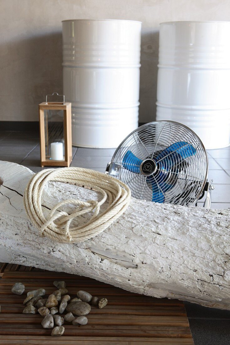 Coiled rope on whitewashed tree trunk on floor in front of fan with white-painted metal drums in background