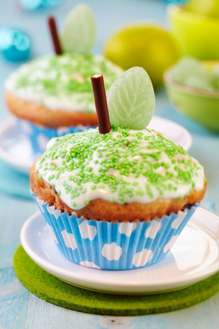 Apple muffins decorated as apples with icing, green sprinkles, chocolate sticks and leaf-shaped sweets