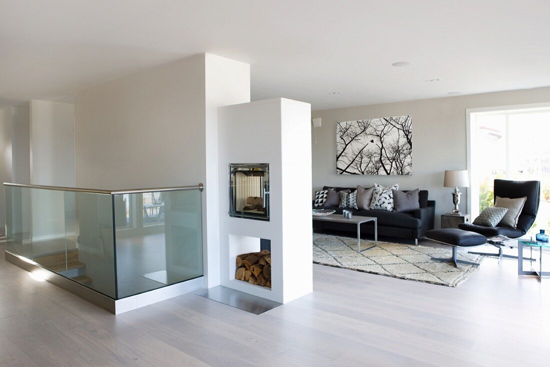 Large interior with dark sofa set and double-sided fireplace in partition; head of staircase with glass balustrade in foreground