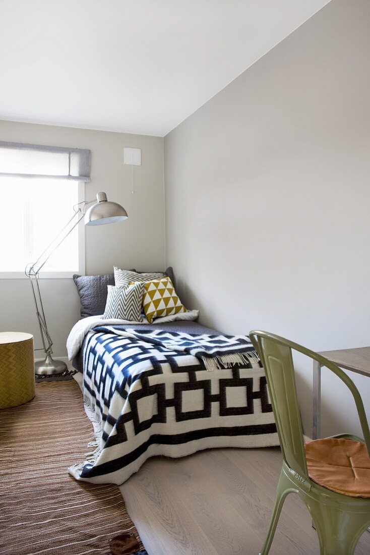 Metal standard lamp over bed with textiles in graphic patterns and olive green, vintage metal chair in foreground chair