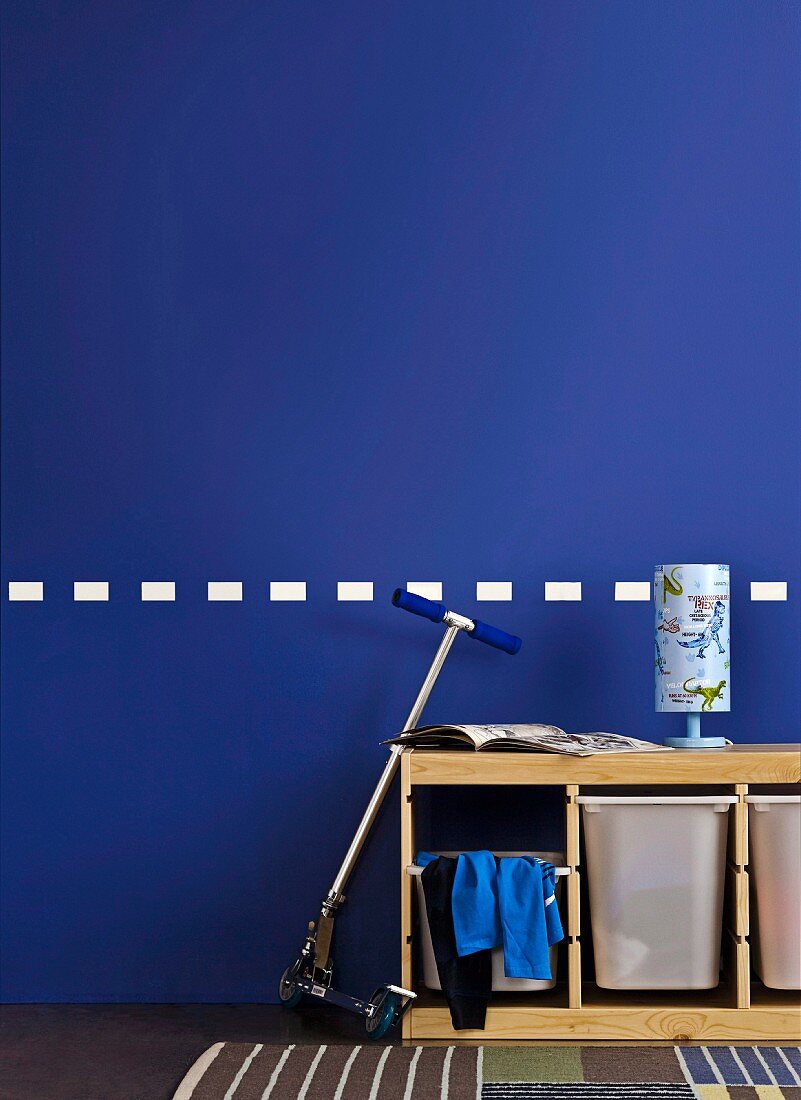 Scooter leaning on practical storage boxes in child's bedroom with dinosaur lamp and blue wall