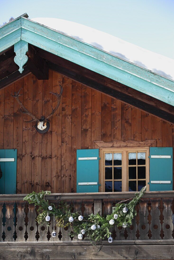 Schnee auf Berghütte mit türkisfarbenen Fensterläden und Ortgangbrett, nostalgische, gestrickte Weihnachtsdeko am rustikalen Balkongeländer