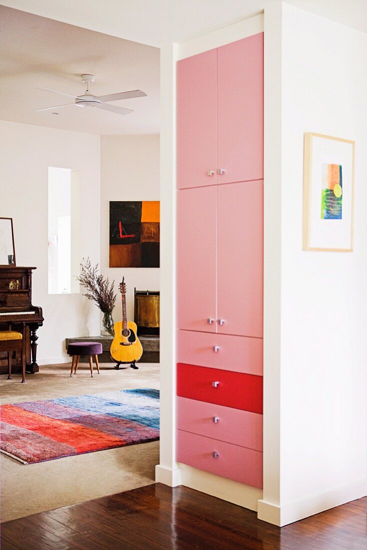 White cupboard with fronts painted pink and red; piano, guitar and brightly striped rug in music area in background