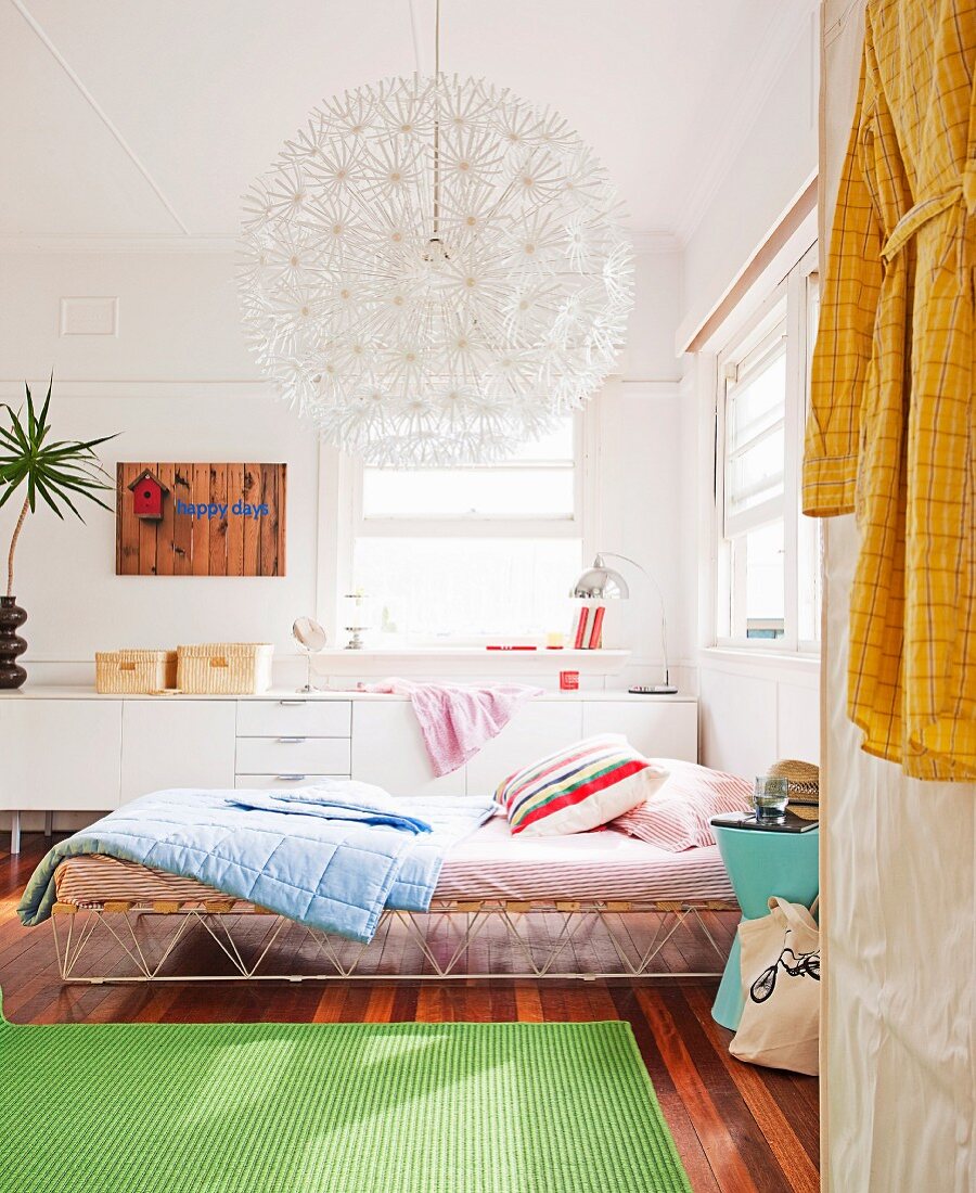Green rug on wooden floor, bed with retro metal frame and scatter cushions, spherical lamp with many flower-shaped elements and white sideboard in background