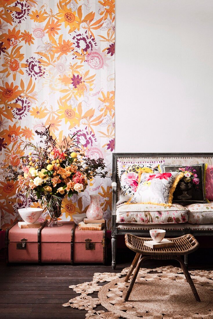 Romantisches Arrangement - Rattan Schemel auf rundem Sisalteppich vor Sitzbank mit Polstern, daneben Blumenstrauss auf antiken, rosa Koffer auf Boden, vor geblümtem Wandbehang
