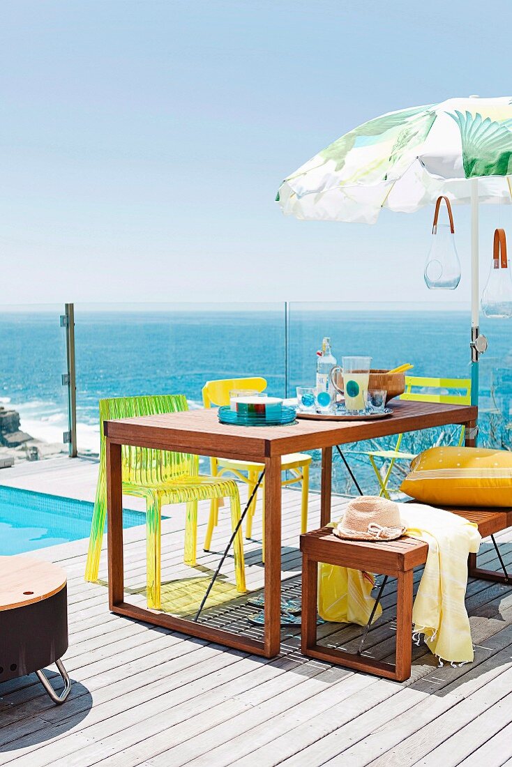 Wooden table, matching bench and yellow chairs on sunny wooden terrace beside pool with sea view through glass balustrade