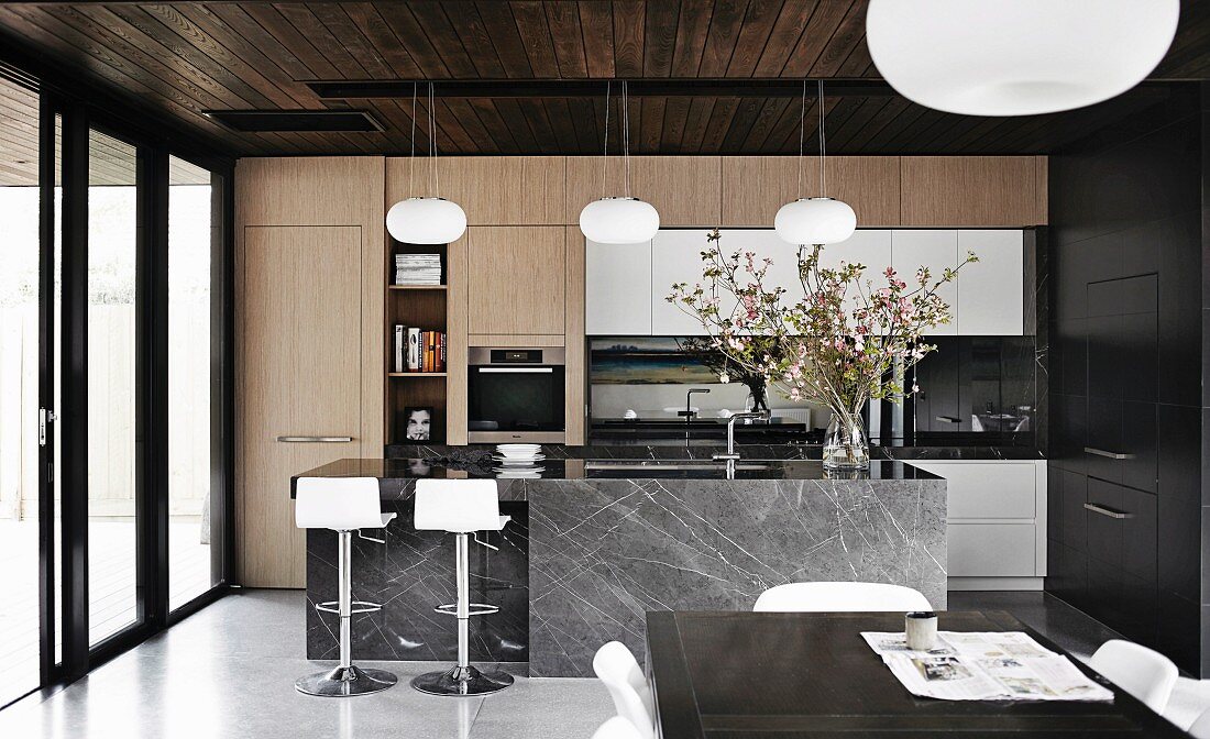 View from dining area into fitted kitchen with pale wooden fronts and bar stools at dark grey stone counter