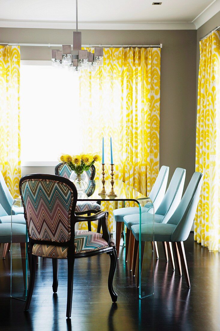 Armchair with patterned upholstery and white-covered chairs at glass table; yellow and white patterned, floor-length curtains on windows beyond