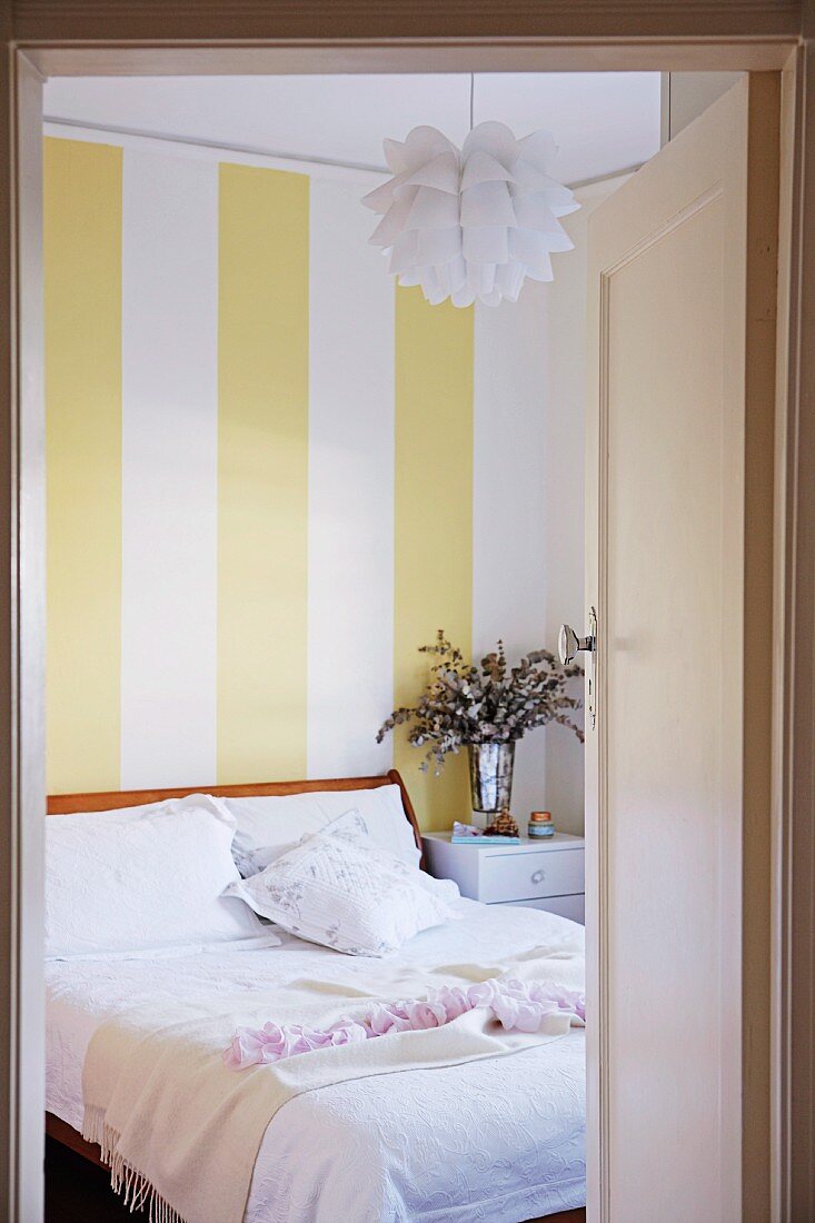 View through open door into bedroom with white bed linen, Bauhaus pendant lamp and yellow and white striped wallpaper