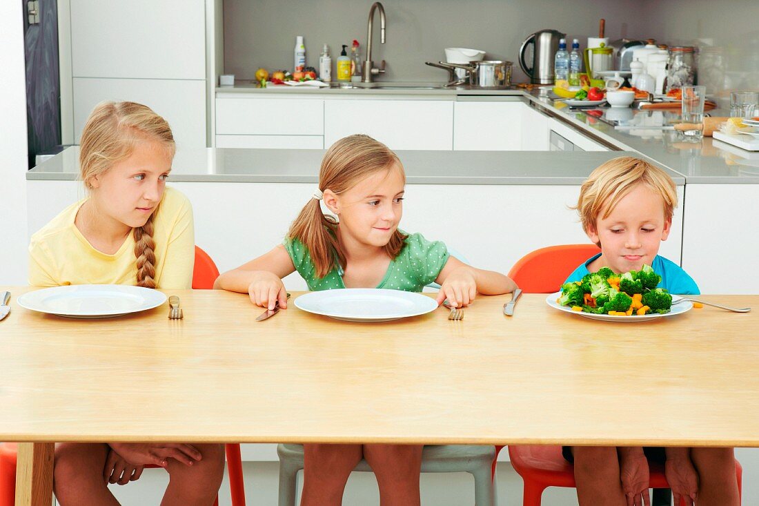 Drei Kinder am Tisch in der Küche starren auf Teller mit Gemüse