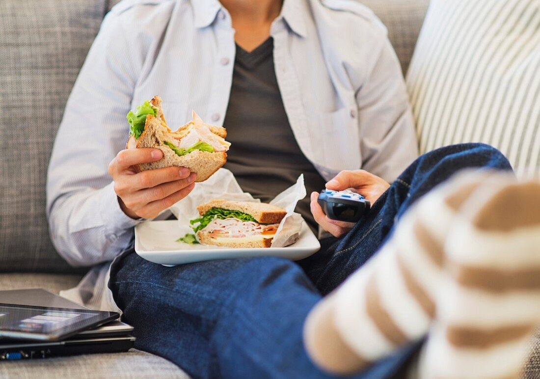 A young man sitting on the sofa holding a sandwich and a remote control