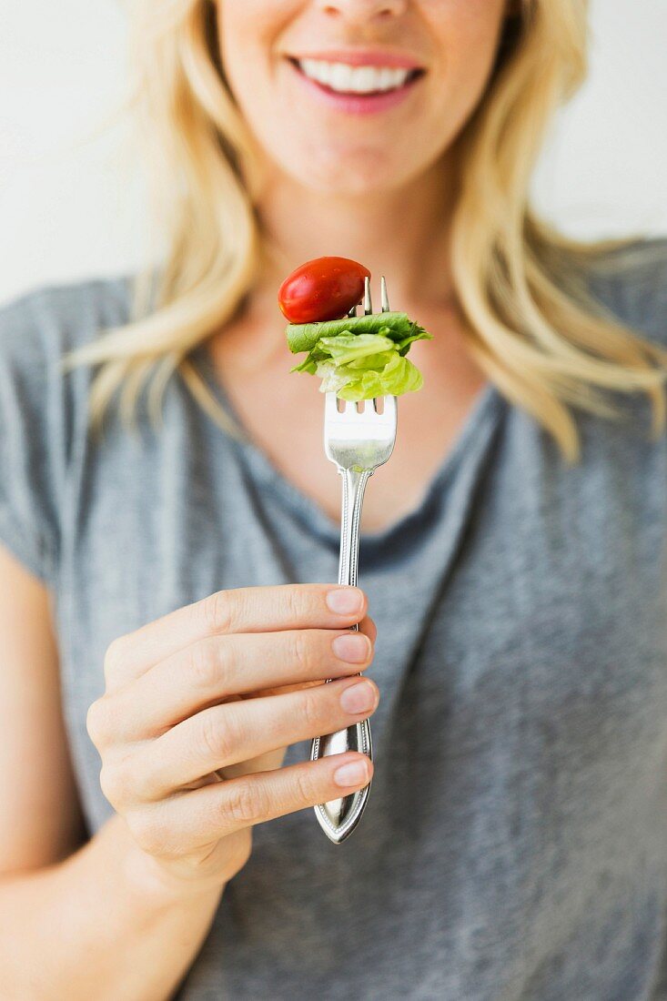 A young woman holding a fork with a tomato and a lettuce leaf skewered on it