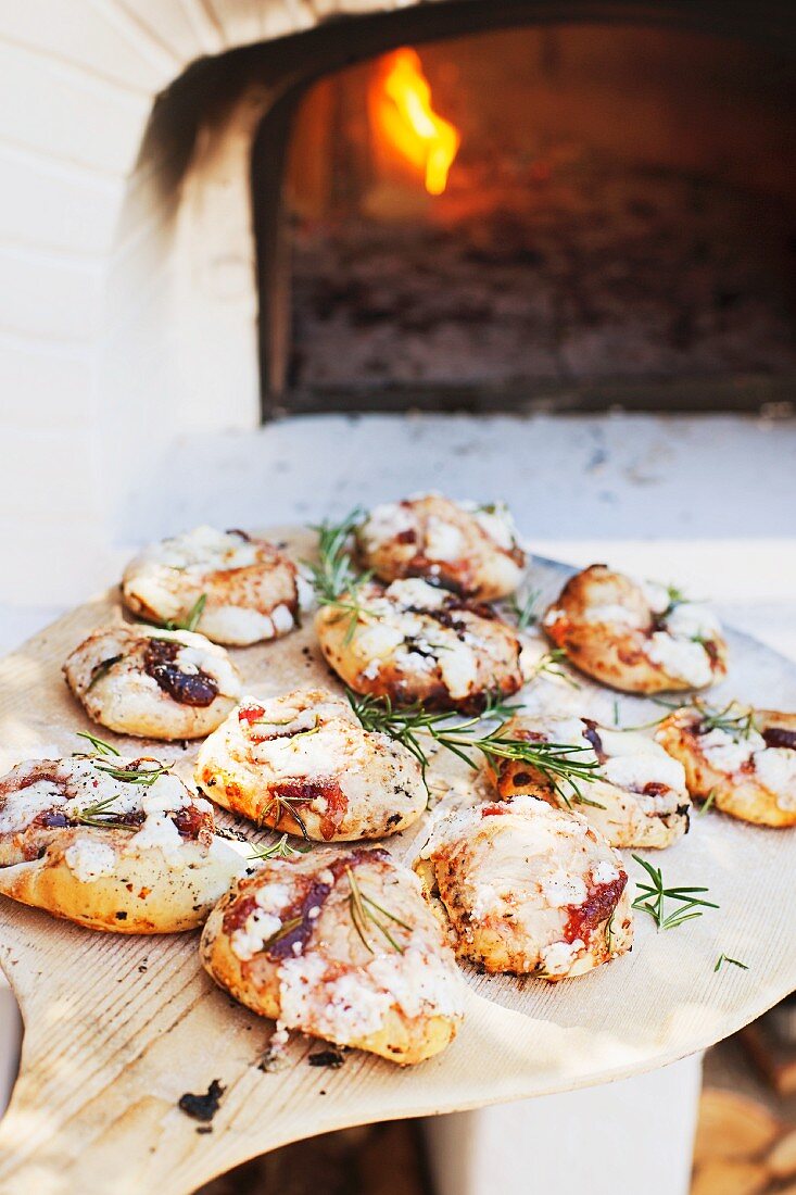 Mini pizzas with tomatoes and rosemary on a pizza paddle in front of a wood-fired oven