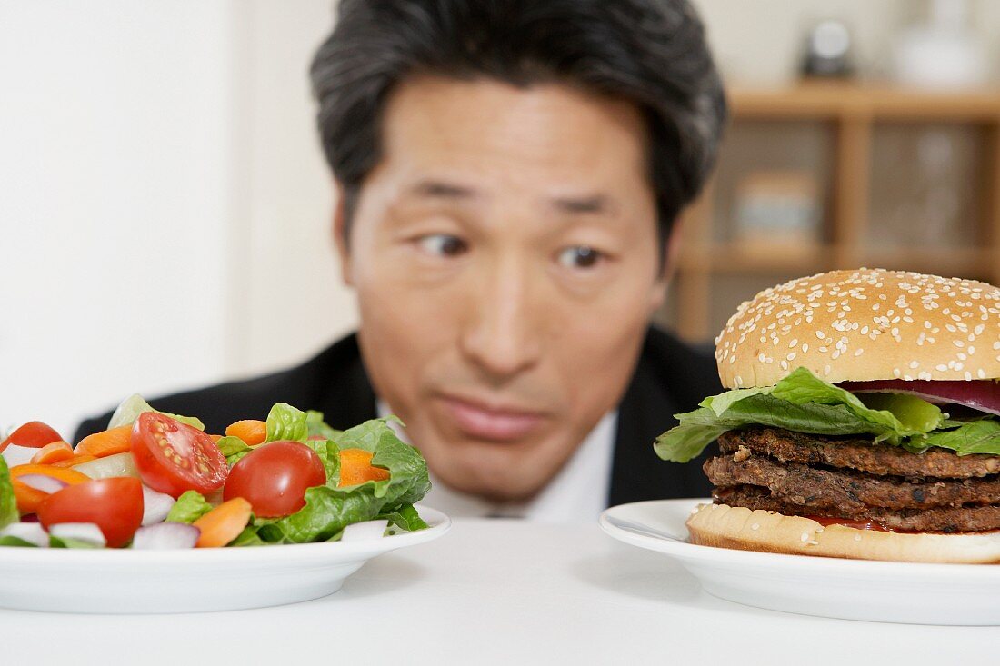 Asiatischer Mann vor Teller mit Salat & Teller mit Hamburger