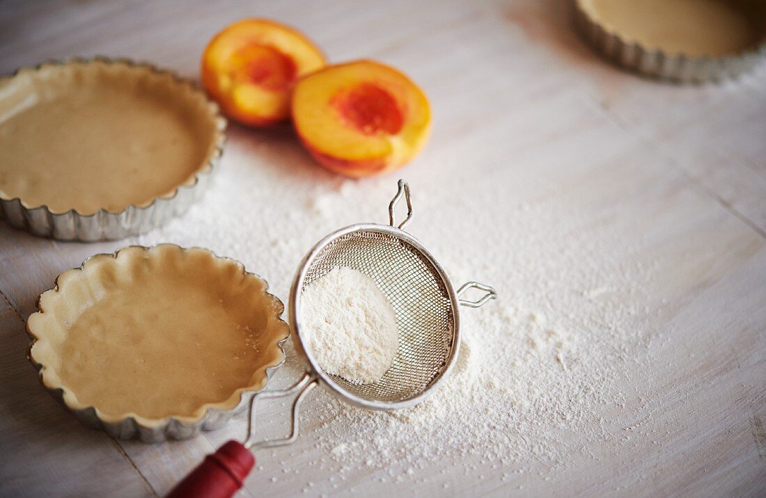Work surface for tart making, including tart pans with pastry, fresh peaches and flour