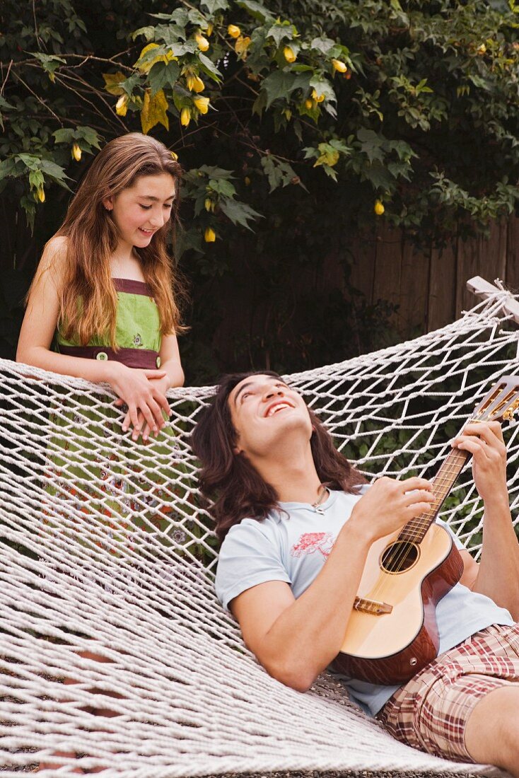 Young man lying in a hammock, playing a ukulele to his sister