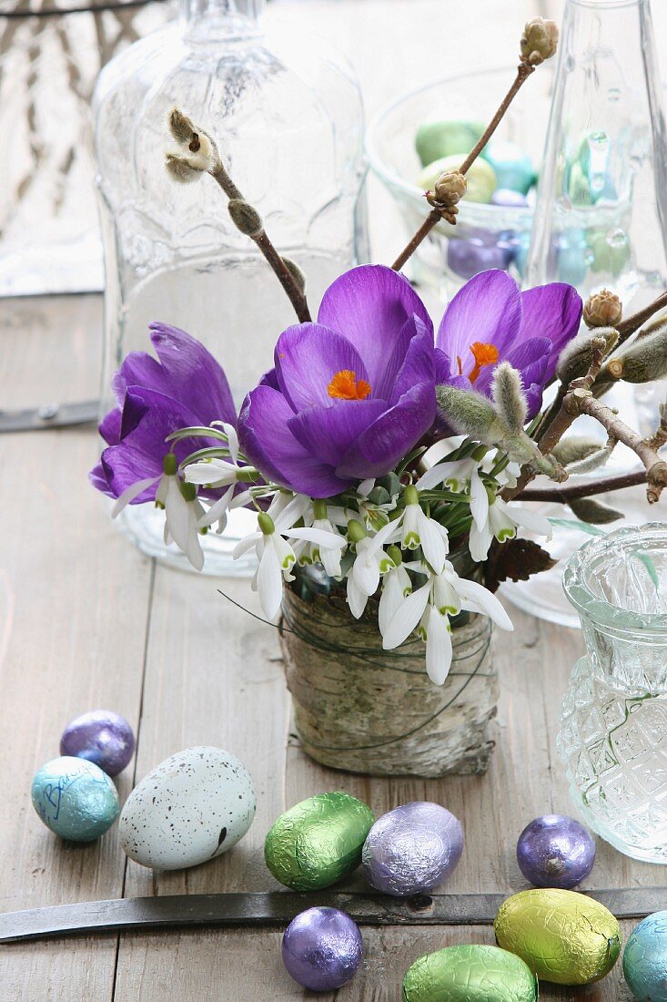 Spring posy of purple crocuses, snowdrops and willow catkins next to chocolate Easter eggs
