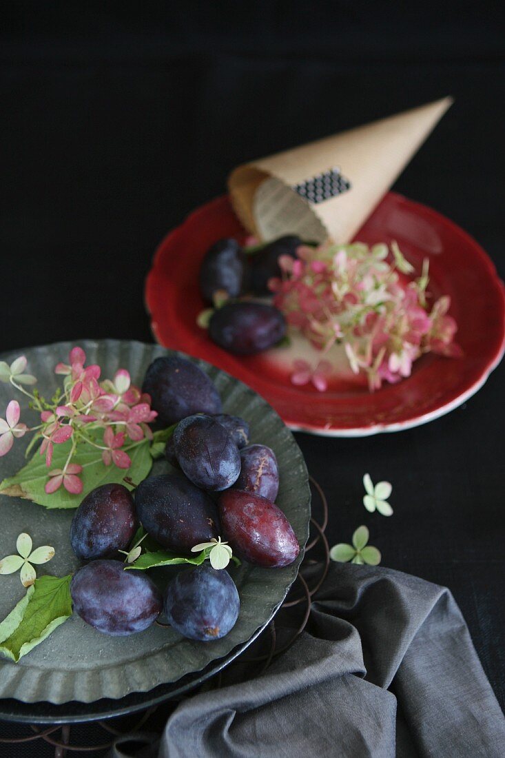 Plums and flowers on tin plates next to a paper cone on a red plate