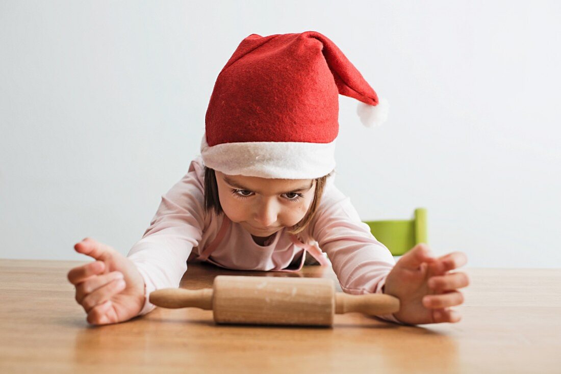 A little girl wearing a Santa hat sitting at a table with a rolling pin