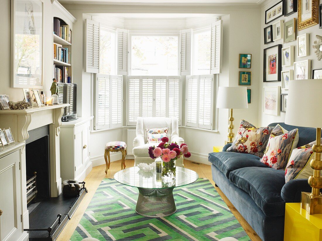 60s table on rug with geometric pattern and floral scatter cushions on sofa; bay window with interior shutters in background