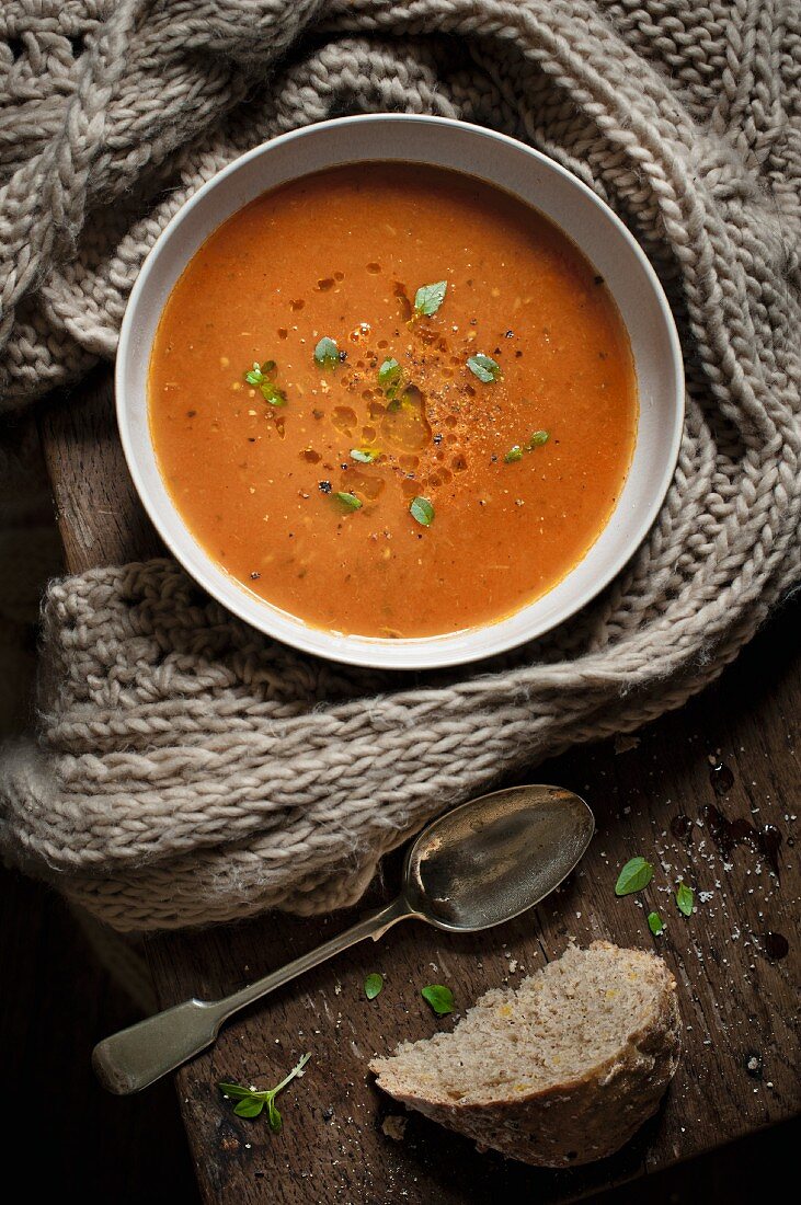 A bowl of tomato soup sprinkled with basil leaves with a knitted scarf, spoon and a slice of bread.
