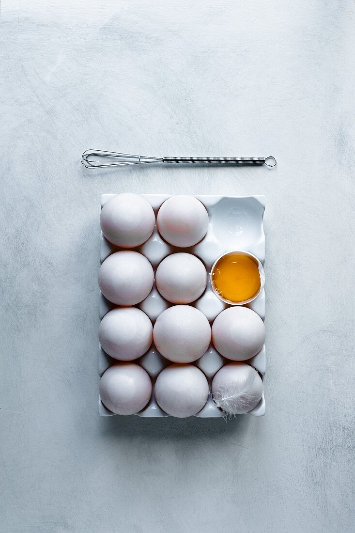 Eier (eines aufgeschlagen) im Eierkarton mit Feder und Schneebesen