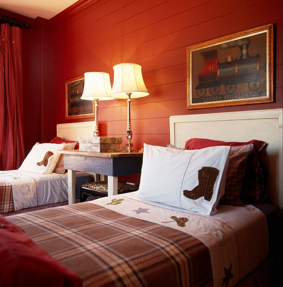 Zwei Landhausbetten mit Karodecken vor rot gestrichener Holzwand und traditionelle Nachttischleuchten mit Messingfuss in einem Schlafzimmer