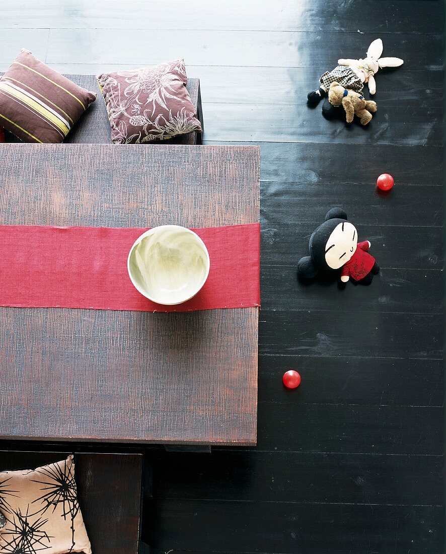 Low, Oriental table decorated with red runner and small cushions on benches; Oriental fabric doll on black-varnished wooden floor