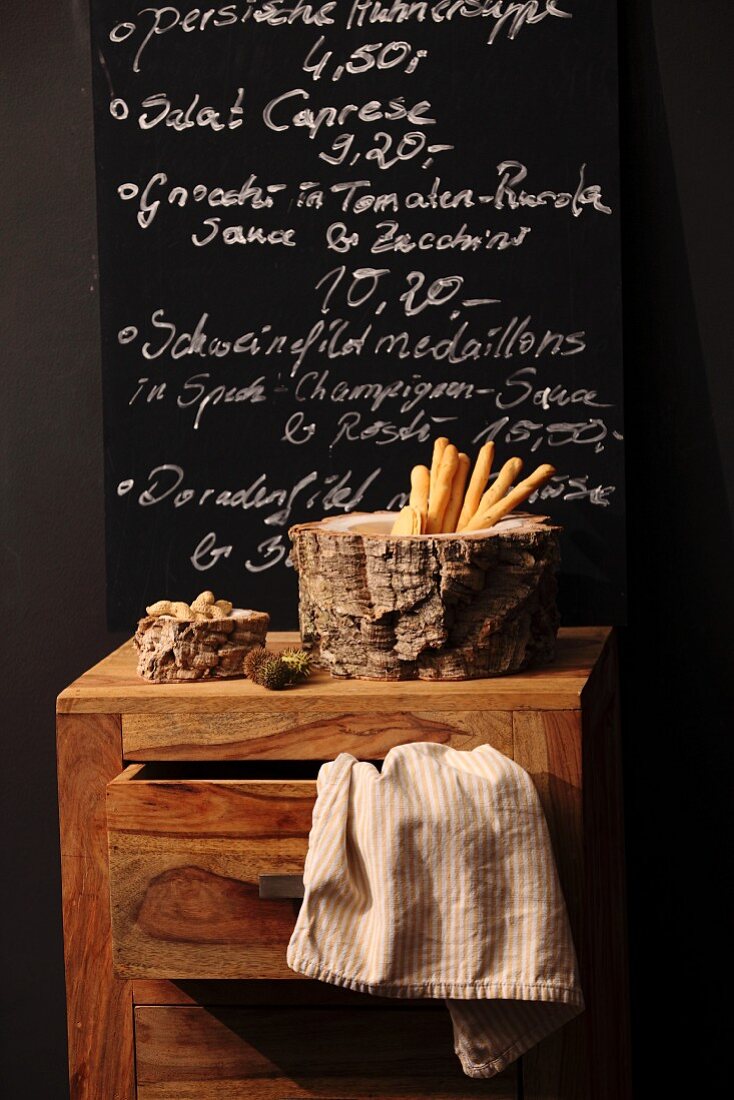 Dishes of nuts and breadsticks surrounded by cork bark on chest of drawers in front of menu on blackboard