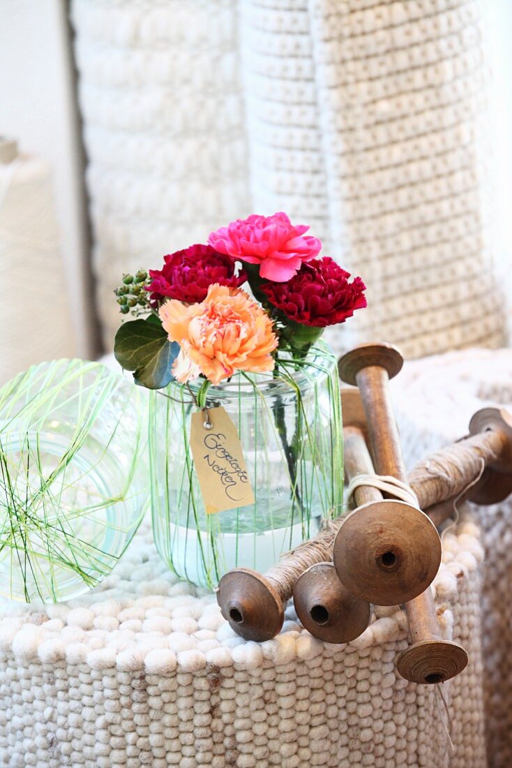 Pinks in preserving jar wrapped in yarn and wooden spindles on upended, rolled wool rug