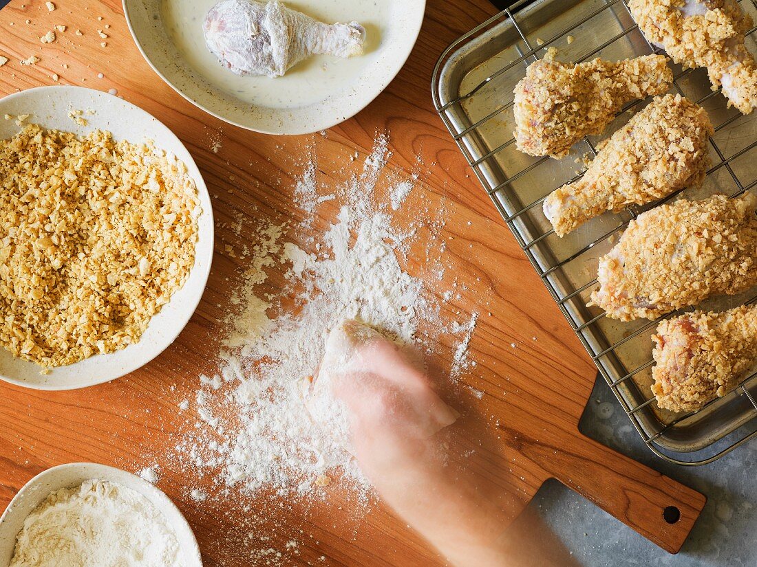 Preparation for breading baked chicken legs in a crushed potato chip crust with an hand in action sprinkling flour