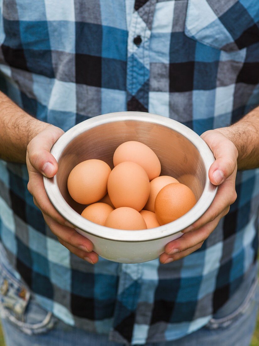 Hands holding a modern bowl of brown eggs with a blue plaid shirt in the background