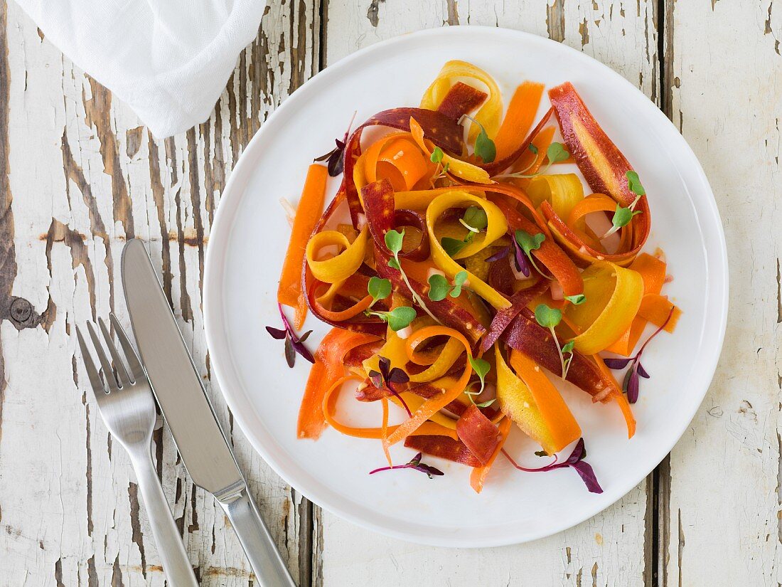 Bright yellow, orange and red carrot salad with micro-greens on a white plate sitting on an antique white wood surface