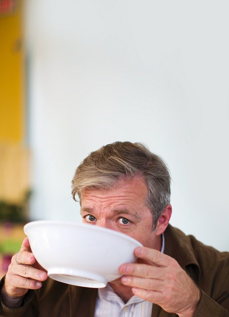 Man looking at the camera while the lower part of his face is covered as he drinks from a white soup bowl