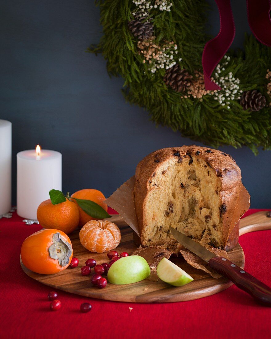 Italian panettone cut open on a cutting board with fruit on a red tablecloth with white candles and a wreath in the background