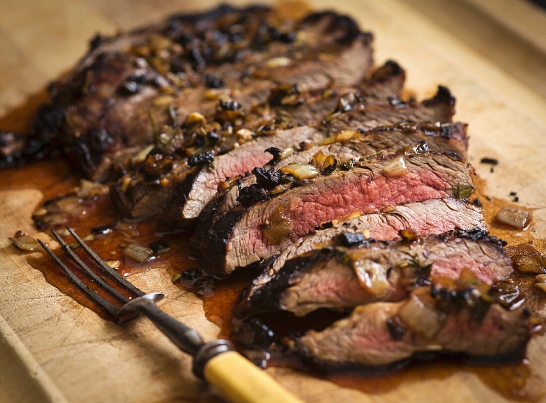 London Broil steak sliced on a cutting board with carmelized onions and a vintage carving fork