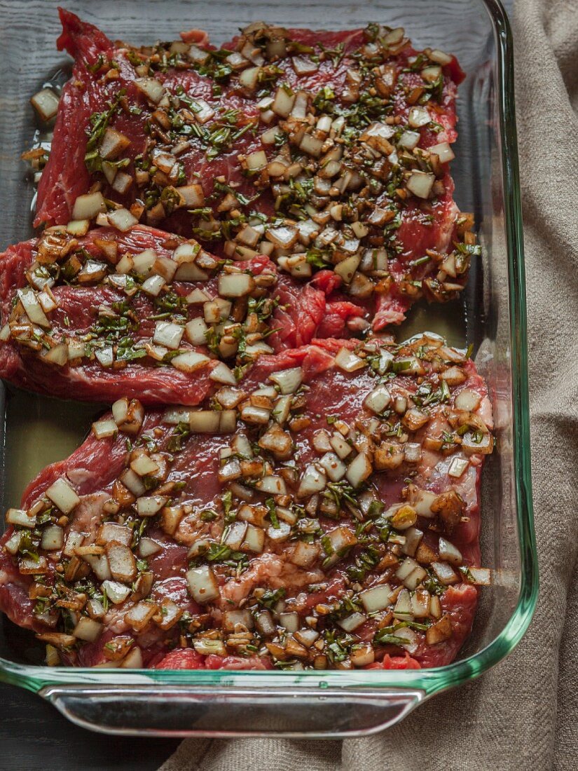 Raw skirt steak marinating in raw chopped onions and herbs in a glass dish